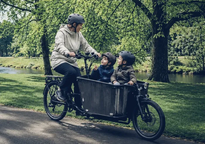 Förälder cyklar en lådcykel med två barn i lådan genom en park en sommardag